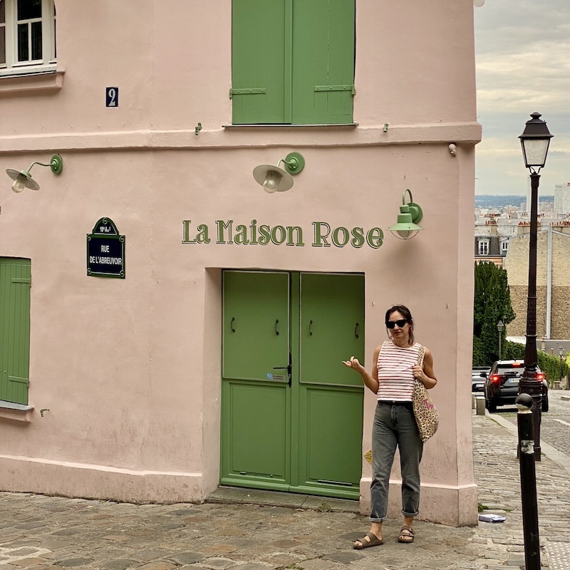 Mónica (El Viaje de Sofi) frente a la fachada rosa del restaurante La Maison Rose en la esquina de la Rue de l'Abreuvoir en Montmartre, París.