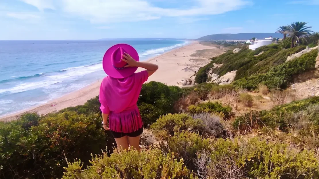 El Viaje de Sofi (Mónica) de espaldas con sombrero fucsia mirando la Playa de Atlanterra en Cádiz desde un acantilado.