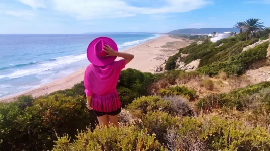El Viaje de Sofi (Mónica) de espaldas con sombrero fucsia mirando la Playa de Atlanterra en Cádiz desde un acantilado.