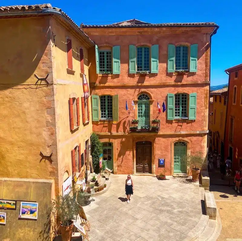 Plaza principal de Roussillon con edificios de color ocre rojizo, ventanas verdes y cielo azul intenso.