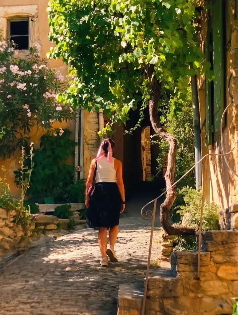 El Viaje de Sofi (mónica) caminando por una calle empedrada de Gordes bajo una parra verde y fachadas de piedra clara.