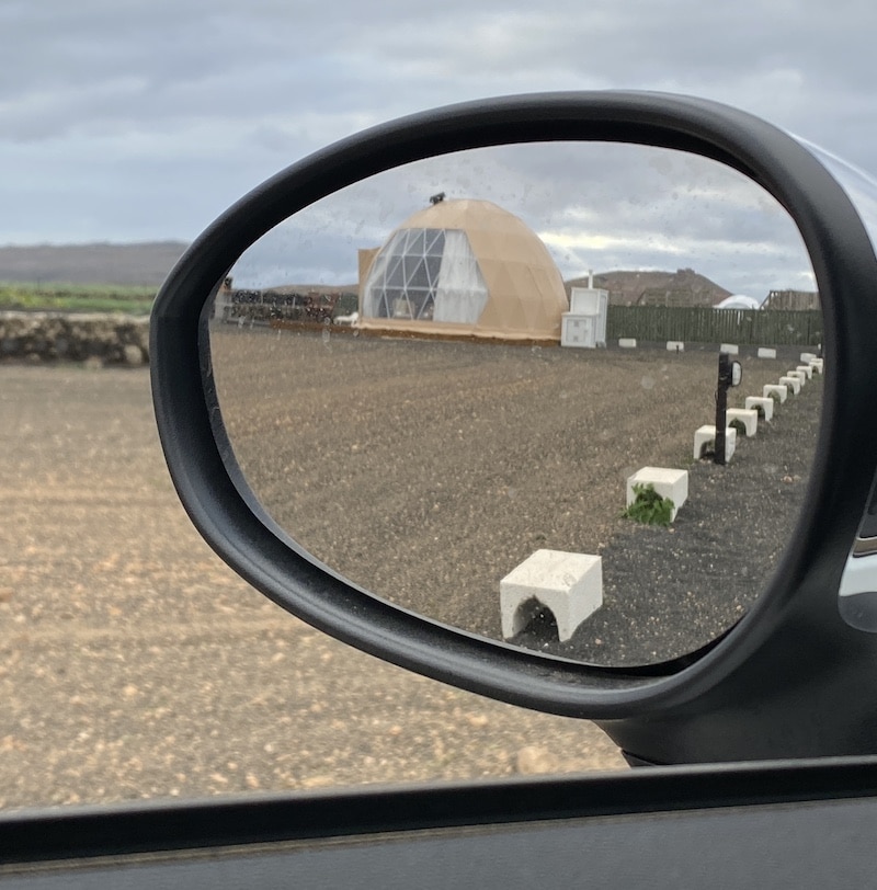 Reflejo en el espejo retrovisor de un alojamiento tipo domo en Teguise con zona de aparcamiento.