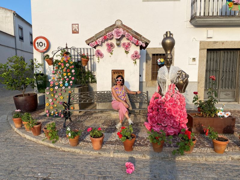 Mónica sentada en un banco junto a una estatua decorada con flores rosas tradicionales en Villanueva del Duque