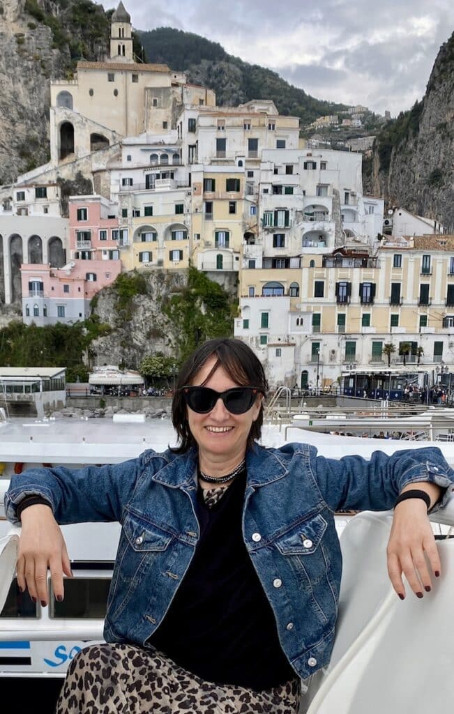 El Viaje de Sofi (Mónica) sentada en la cubierta de un ferry y las vistas del pueblo y acatantilados de Amalfi detrás.