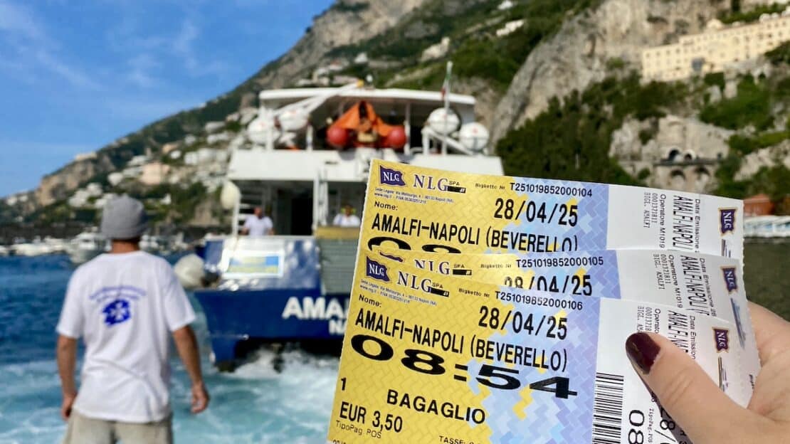 Mano sosteniendo billetes de ferry hacia Nápoles con el mar y los acantilados de fondo.